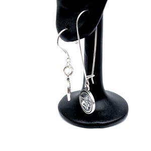 May include: A pair of silver-coloured drop earrings. One earring has a small charm, the other an oval pendant with a detailed image. The earrings are displayed on a black earring stand against a white background.