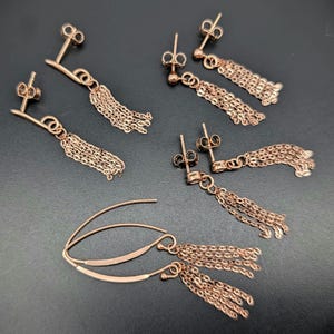 May include: Several pairs of rose gold earrings, including stud and drop styles. The earrings have fine chain tassels and post or hook fastenings. The earrings are presented on a dark surface, highlighting their detailed design and metallic lustre.