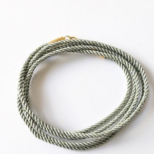 Può includere: Una collana a corda color argento avvolta su se stessa con una trama intrecciata. La collana è fissata con piccoli terminali in metallo dorato lucido e una chiusura abbinata, creando un anello circolare continuo di corda grigio metallizzato.