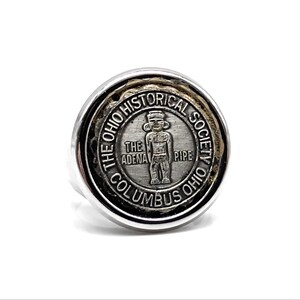 May include: A circular silver-toned ring featuring a central medallion with a relief of a standing figure. The text reads THE OHIO HISTORICAL SOCIETY COLUMBUS OHIO and THE ADENA PIPE. The design is set within a polished metal frame with a dark textured border.
