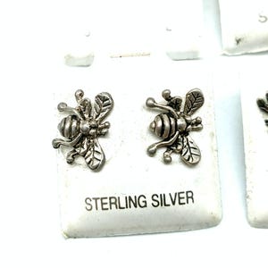 May include: A pair of sterling silver bee stud earrings attached to a white display card with the text STERLING SILVER printed in black. The earrings feature a detailed insect design with textured wings and a segmented body in a metallic finish.