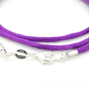 May include: Close-up of a purple cord necklace with a silver clasp. The necklace features a bright purple cord and a shiny silver clasp with a small tag that reads "025". The clasp is open, showing the details of the mechanism.