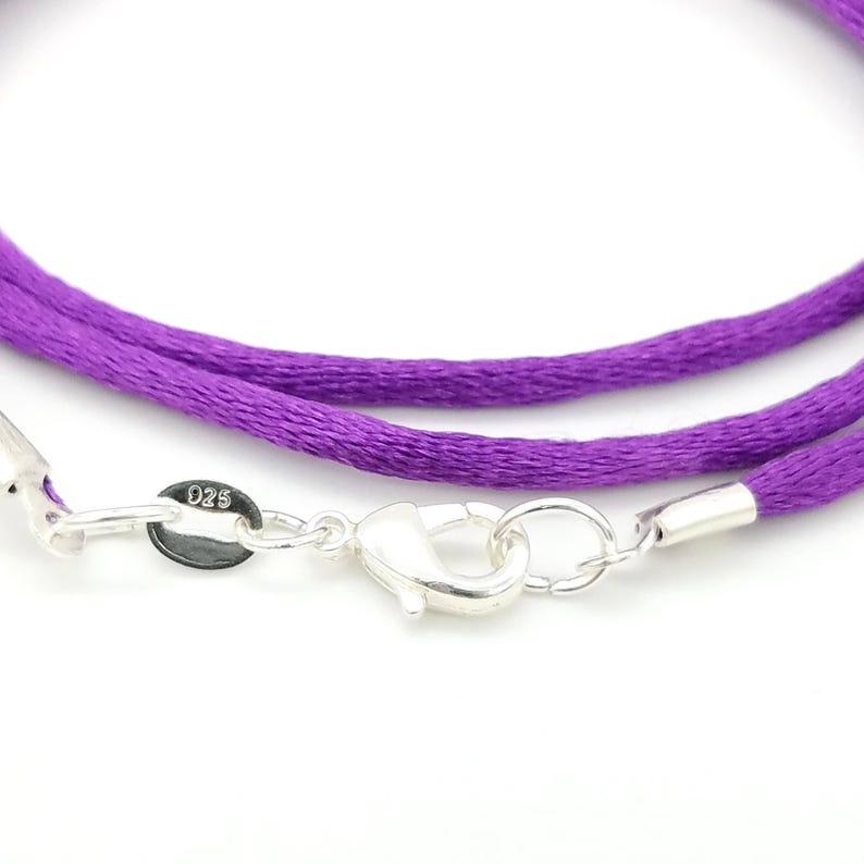 May include: Close-up of a purple cord necklace with a silver clasp. The necklace features a bright purple cord and a shiny silver clasp with a small tag that reads "025". The clasp is open, showing the details of the mechanism.