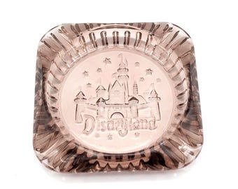 May include: A square, clear ashtray with a pinkish hue. The center features an embossed image of a castle with the word "Disneyland" below. The ashtray has a ridged design around the edges.