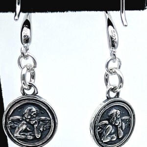 May include: A pair of silver drop earrings. Each earring has a circular pendant with a detailed relief of an angel. The earrings are connected by small silver rings and have a hook closure. The metal is stamped with "S925".