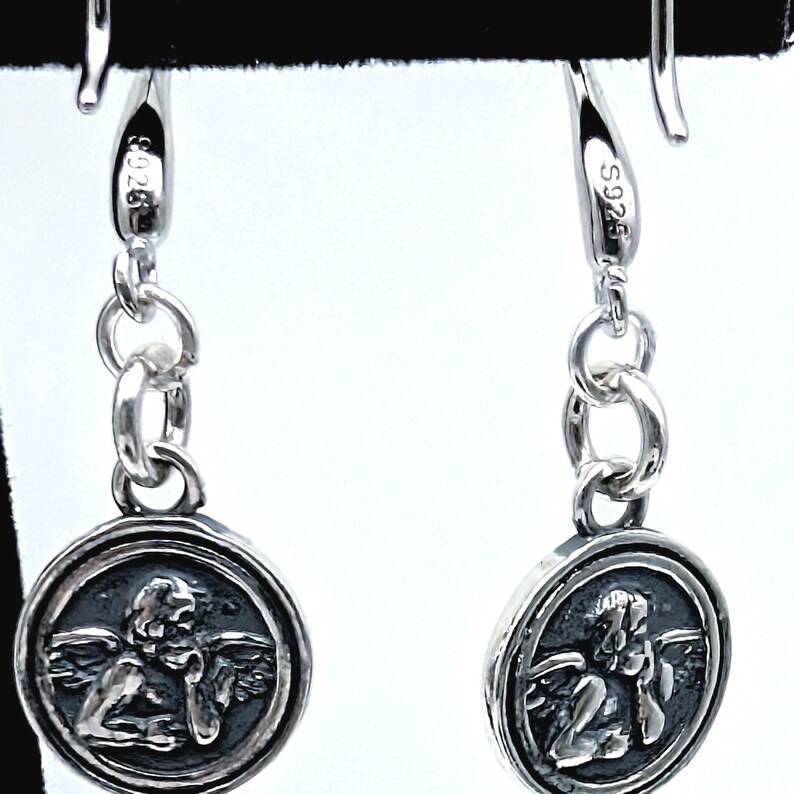 May include: A pair of silver drop earrings. Each earring has a circular pendant with a detailed relief of an angel. The earrings are connected by small silver rings and have a hook closure. The metal is stamped with "S925".