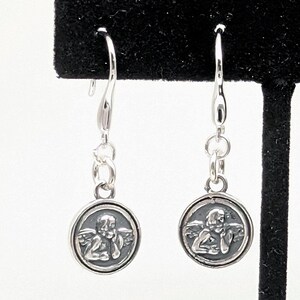 May include: A pair of silver drop earrings. Each earring has a small, circular charm with a detailed relief of an angel. The charms hang from a curved hook with small connecting rings.