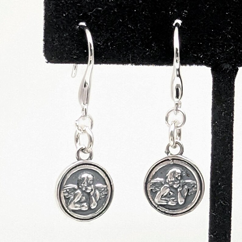 May include: A pair of silver drop earrings. Each earring has a small, circular charm with a detailed relief of an angel. The charms hang from a curved hook with small connecting rings.