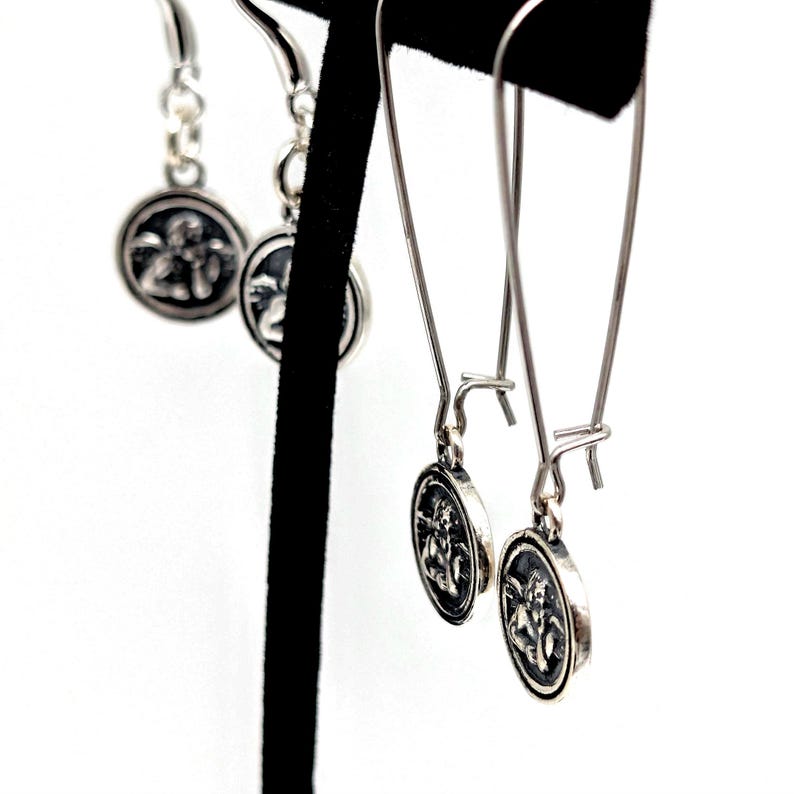 May include: Silver earrings with a circular pendant featuring a detailed relief design. The earrings are displayed on a black stand against a white background. The pendant design appears to be a classical figure.