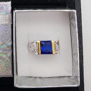 May include: A silver ring with a large, square, deep blue gemstone at the center, flanked by smaller clear stones and gold-colored accents. The ring is presented in a white-lined box, with a decorative silver and iridescent gift box visible.