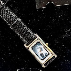Pu&ograve; includere: Un orologio da polso rettangolare con cassa argentata e cinturino in pelle nera. Il quadrante dell'orologio presenta un'immagine scura. Il cinturino &egrave; strutturato e ha una fibbia argentata. L'orologio &egrave; esposto su una superficie nera.