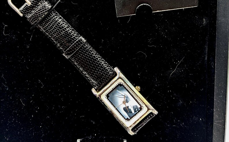 Pu&ograve; includere: Un orologio da polso rettangolare con cassa argentata e cinturino in pelle nera. Il quadrante dell'orologio presenta un'immagine scura. Il cinturino &egrave; strutturato e ha una fibbia argentata. L'orologio &egrave; esposto su una superficie nera.