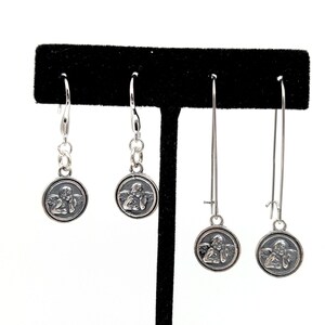 May include: Four pairs of silver earrings. Each earring has a circular pendant with a detailed relief of an angel. Two pairs have a shorter drop with chain links, and two pairs have a longer, linear design.