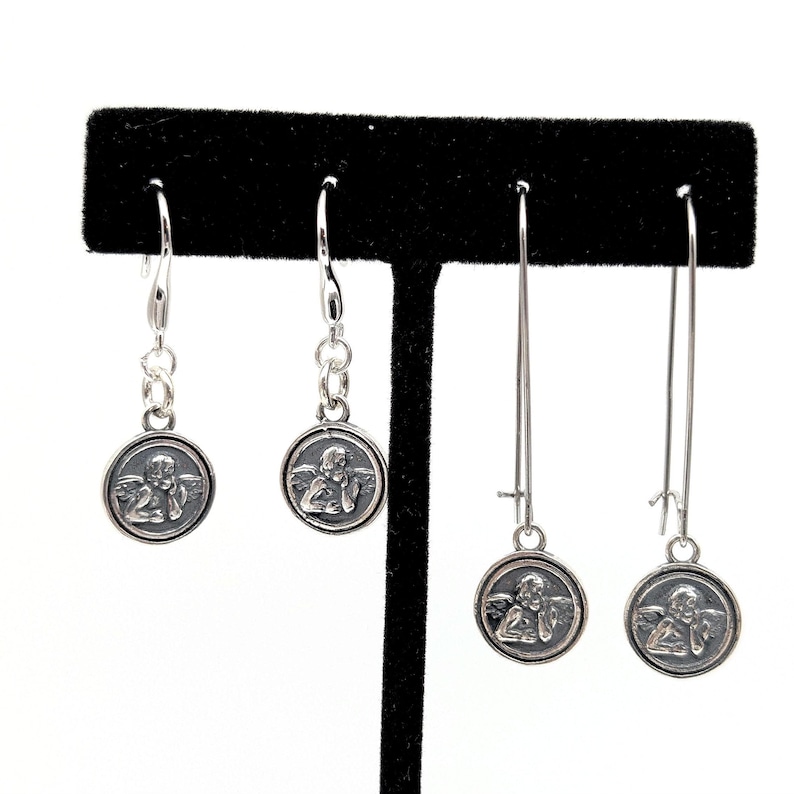 May include: Four pairs of silver earrings. Each earring has a circular pendant with a detailed relief of an angel. Two pairs have a shorter drop with chain links, and two pairs have a longer, linear design.