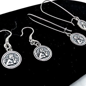 May include: Silver earrings with a cherub design on a small, round pendant. The earrings have a hook and dangle style. The earrings are displayed on a black surface. The design is simple and elegant.