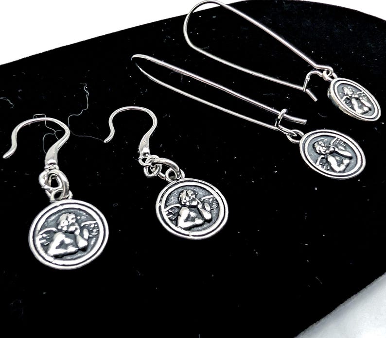 May include: Silver earrings with a cherub design on a small, round pendant. The earrings have a hook and dangle style. The earrings are displayed on a black surface. The design is simple and elegant.