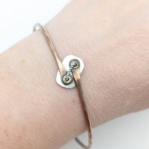 May include: A silver and copper bracelet with a unique, twisted design. The bracelet features a circular, silver charm with a copper accent.