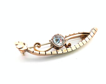 May include: A gold-toned brooch shaped like a canoe, featuring a central round, light blue faceted gemstone. The metal frame has a segmented, ribbed design along the curved edge, with a small leaf-shaped detail at one end and scrollwork accents.