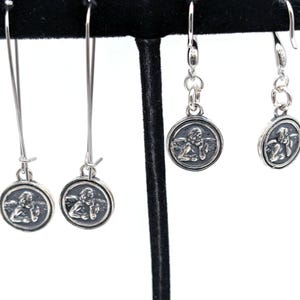 May include: Silver earrings with a cherub design in a circular frame. The earrings are displayed on a black stand, highlighting the intricate details. The image showcases the delicate design and craftsmanship of the jewellery, emphasizing the cherub's detailed features.