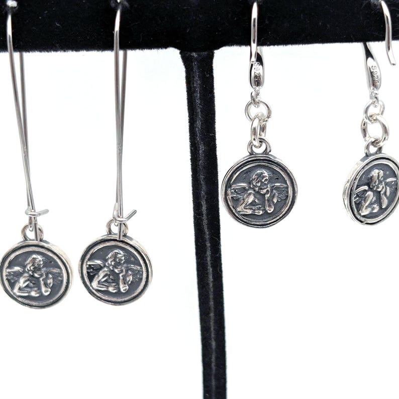 May include: Silver earrings with a cherub design in a circular frame. The earrings are displayed on a black stand, highlighting the intricate details. The image showcases the delicate design and craftsmanship of the jewellery, emphasizing the cherub's detailed features.