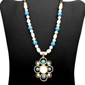 May include: A gold-toned necklace with a pendant and beaded chain. The chain features white and blue beads. The pendant has a white center, surrounded by gold filigree and blue accents. The necklace is displayed on a black mannequin.