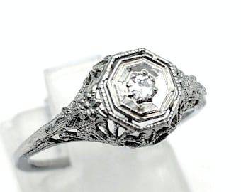 May include: A silver-toned ring featuring an octagonal setting with a central round clear stone. The band and the setting display intricate, openwork filigree metal detailing throughout, creating a textured and ornate appearance on the metal surface.