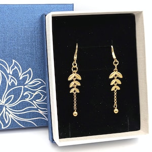 May include: A pair of gold-colored leaf earrings displayed in a white box with a black interior. The earrings have a delicate design with multiple leaf shapes and a small dangling ball at the bottom. The box is inside a blue decorative box.