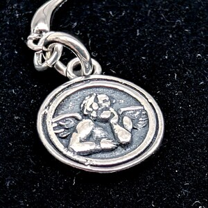 May include: A silver pendant with a detailed relief of a cherub inside a circular frame. The cherub has wings and is touching its face. The pendant is attached to a silver chain.