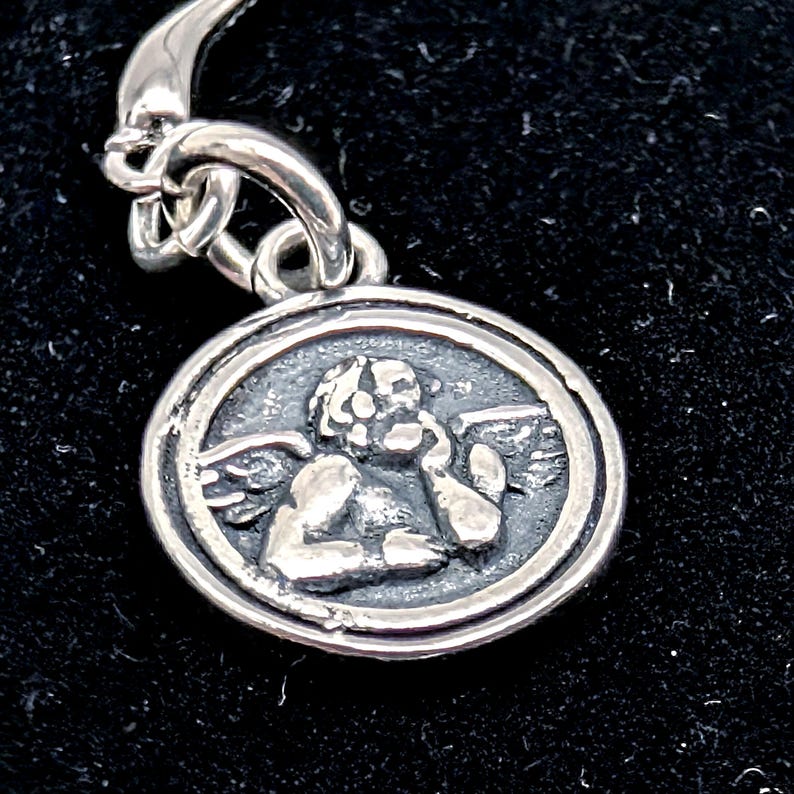 May include: A silver pendant with a detailed relief of a cherub inside a circular frame. The cherub has wings and is touching its face. The pendant is attached to a silver chain.