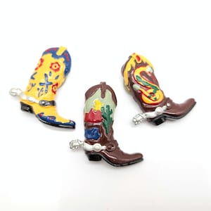 May include: Three miniature cowboy boots in yellow, brown, and red with silver spurs. The boots are decorated with floral and desert-themed designs. The boots are arranged on a white surface.