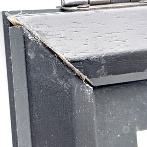 Pu&ograve; includere: Primo piano di un angolo di cornice in legno nero con una cerniera argentata. La cornice ha una finitura invecchiata con usura visibile. L'angolo mostra una separazione nel legno.