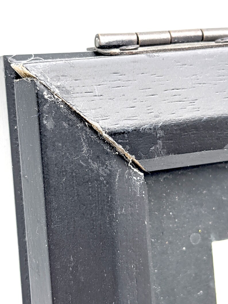Pu&ograve; includere: Primo piano di un angolo di cornice in legno nero con una cerniera argentata. La cornice ha una finitura invecchiata con usura visibile. L'angolo mostra una separazione nel legno.