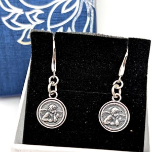 May include: Silver-tone dangle earrings. Each earring has a small, round pendant with a detailed relief of a cherub. The earrings are presented on a black surface, with a blue and white patterned box in the background.