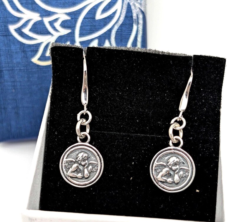 May include: Silver-tone dangle earrings. Each earring has a small, round pendant with a detailed relief of a cherub. The earrings are presented on a black surface, with a blue and white patterned box in the background.