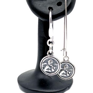 May include: Silver dangle earrings with a circular pendant depicting an angel. The earrings have a hook fastening and a long, slender drop. The angel is shown with wings and a calm expression. The earrings are displayed on a black earring stand.