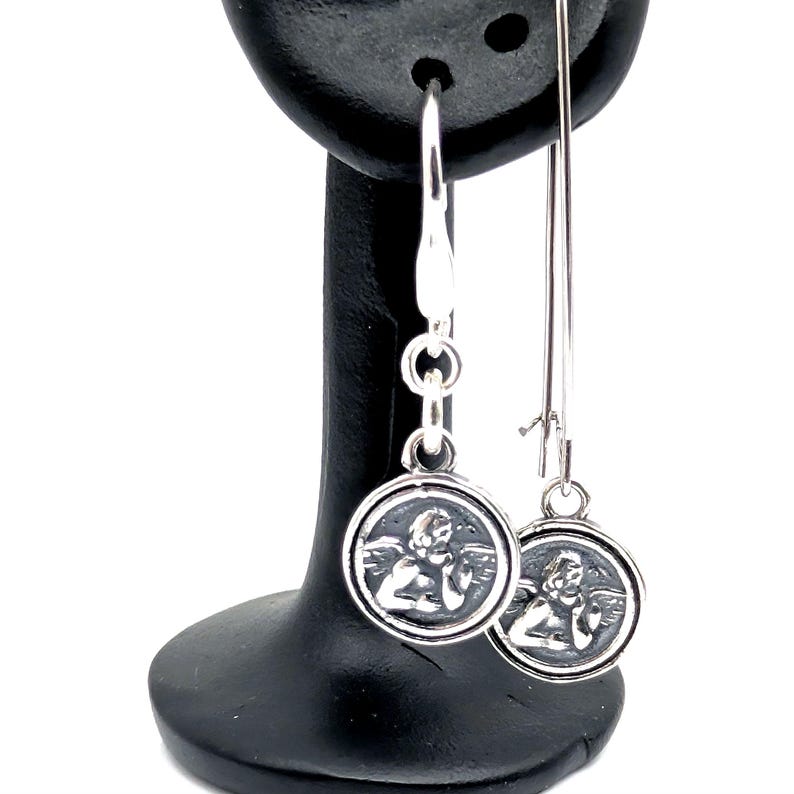May include: Silver dangle earrings with a circular pendant depicting an angel. The earrings have a hook fastening and a long, slender drop. The angel is shown with wings and a calm expression. The earrings are displayed on a black earring stand.
