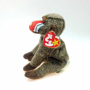 May include: A brown and tan plush baboon toy with a red, white, and blue nose. The toy has brown feet and ears. A red heart-shaped tag with gold trim and text is attached to the baboon's ear.