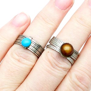 May include: Two silver rings, each with a central stone. One ring features a bright turquoise stone, while the other has a brown, eye-like stone. The rings are worn on fingers, showcasing their design.