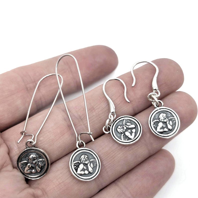 May include: Silver earrings with a circular pendant depicting an angel. The earrings have a hook fastening and a long, slender drop. The angel pendant is detailed with a classical design.