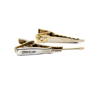 Può includere: Due fermacravatte vintage. Uno è a forma di cacciavite con la scritta "Snap-on" sul manico. L'altro è una sega color oro. Entrambe le clip hanno una chiusura color oro.