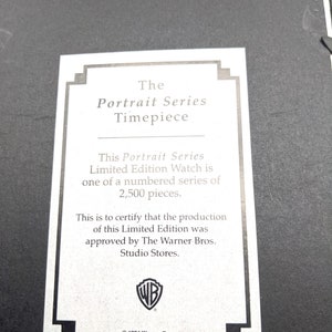 Pu&ograve; includere: Una carta bianca con testo nero, con la scritta "The Portrait Series Timepiece". La carta descrive un orologio in edizione limitata di 2.500 pezzi, approvato da The Warner Bros. Studio Stores. Il logo Warner Bros. &egrave; visibile.