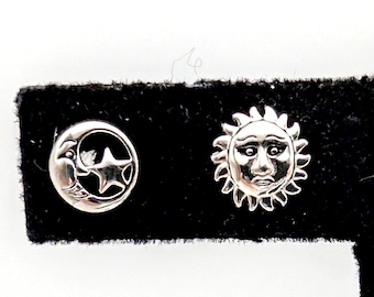 May include: Silver-toned celestial stud earrings. One earring features a crescent moon with a star, the other a smiling sun face. The earrings are displayed on a black surface.