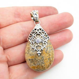 Puede incluir: Un colgante en forma de lágrima con un diseño de filigrana plateada y una pequeña gema púrpura. El colgante presenta una piedra natural con tonos marrones y beige. El engaste plateado tiene un diseño calado ornamentado.