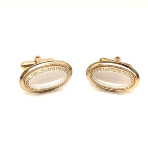 May include: A pair of gold-toned cufflinks with an oval, white center. The gold border features a repeating geometric pattern. The cufflinks have a classic design and are set against a white background.