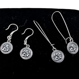 May include: Four silver earrings featuring cherub designs. Two pairs are shown, each with a different hook style. The cherub is within a circular frame. The earrings are presented on a black surface.