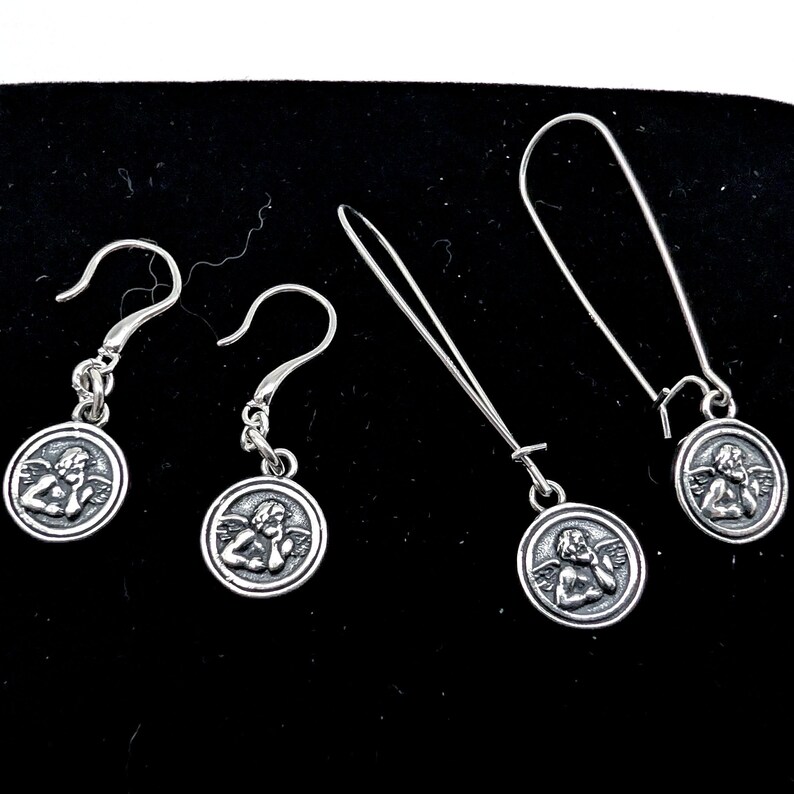 May include: Four silver earrings featuring cherub designs. Two pairs are shown, each with a different hook style. The cherub is within a circular frame. The earrings are presented on a black surface.
