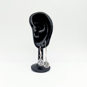 May include: Silver dangle earrings with a circular charm featuring a detailed design. The earrings are displayed on a black ear-shaped stand against a white background. The charms have a detailed design.
