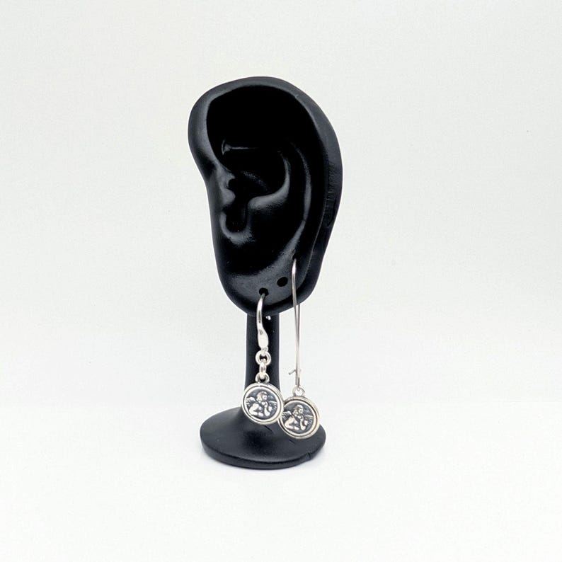May include: Silver dangle earrings with a circular charm featuring a detailed design. The earrings are displayed on a black ear-shaped stand against a white background. The charms have a detailed design.