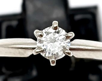 Könnte beinhalten: Ein silberfarbener Ring mit einem einzelnen runden, klaren, facettierten Stein in einer Sechs-Krappen-Fassung. Die Ringschiene ist glatt und verjüngt sich leicht zur zentralen Fassung hin, die den Stein im Brillantschliff sicher hält.