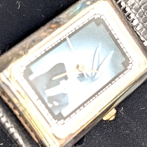 Pu&ograve; includere: Orologio da polso rettangolare argentato con quadrante azzurro e cinturino in pelle nera. L'orologio ha lancette color oro e una corona color oro. Il quadrante ha un design decorativo. Il cinturino ha un motivo strutturato.
